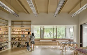 People standing in a modern library interior with wooden shelves and tables, large windows, and a view of greenery outside.