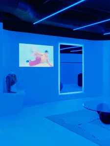 A minimalist installation with blue lighting, a mirror, and a projected image of a person on a screen.