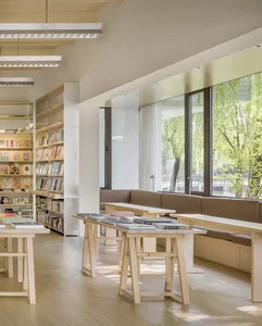 A minimalist library interior with wooden tables, bookshelves, and seating areas near large windows.