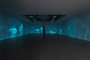 A person standing in a dark room with glowing blue digital projections on the walls creating an immersive minimalist art expe