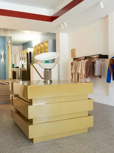 Modern clothing store with brass furniture, featuring a gold pedestal display and clothing racks.