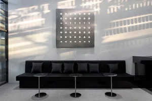 A minimalist office space featuring black furniture, metallic columns, and a metallic panel with a grid of letters.