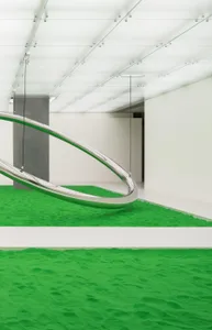 A modern art installation featuring a sleek, curved metal sculpture on a bed of vibrant green sand.