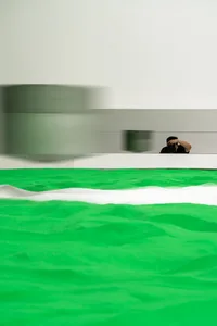 Abstract art installation with green and white sand, white wall, and blurred reflection of photographer.