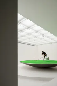A minimalist art installation featuring a green surface being raked within a metallic bowl.