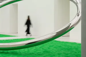 A modern art installation featuring a large, curved metallic structure and green sand floor.