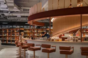 A modern retail bar with a curved wooden counter, leather stools, and a variety of bottles on display.