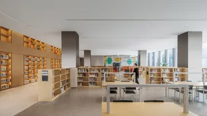 A minimalist library with tall, wooden bookshelves, rows of tables and chairs, and large windows letting in natural light.