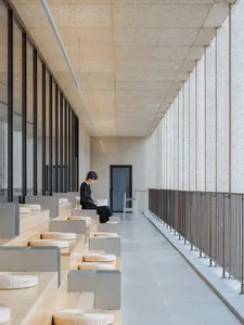 A modern interior corridor with concrete walls, benches, and a person reading a newspaper.