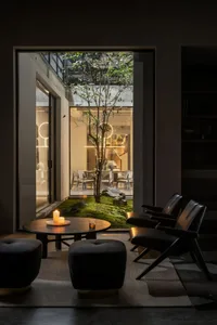 A minimalist living room with wooden furniture, a small table with candles and a drink, and a view of a garden.