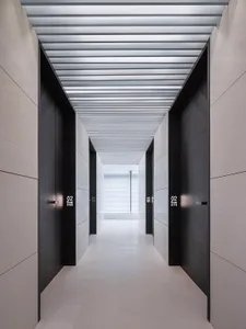 Modern minimalist corridor with white concrete walls and floor, black doors, and a metallic ceiling.