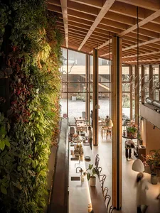 Modern industrial restaurant interior with greenery walls, wooden ceiling beams, and glass windows allowing natural light.