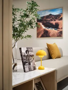 A modern living room featuring a sleek yellow table lamp, a green plant in a vase, and a book titled 'Vivian Maier: A Photogr