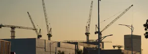 Construction site with multiple cranes and streetlights in the evening light, showing industrial and urban elements.