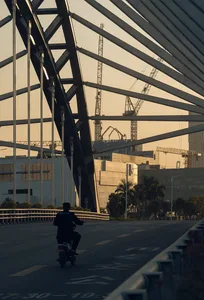 夕陽下，一人騎乘機車穿過鋼構桁架的工地現場。