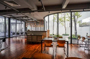 An industrial-style coffee shop with wooden accents, featuring large windows, a bookshelf, and wooden tables and chairs.