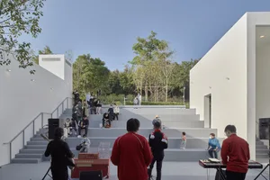 People sitting on concrete steps surrounded by white walls and glass partitions in an urban outdoor pavilion.