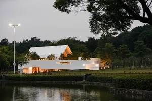 A serene outdoor view of a modern white concrete building with warm indoor lighting and a reflective pond.