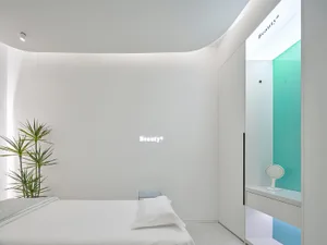 A minimalist spa suite with white walls and furnishings, a green-tinted glass shower, and a neon sign that reads 'Beauty'.