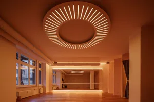 An image of a modern yoga studio with wooden floors and glass walls, featuring a circular light fixture on the ceiling.