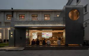 Group of people seated at tables in a modern, industrial-style restaurant exterior with concrete walls and a glass window dis