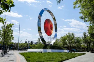 A large metallic circular sculpture with a red glass center, set in a park-like setting with greenery and pathways.