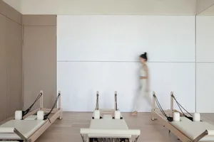 A minimalist fitness studio with three reformer beds aligned against a white wall, and a blurred figure walking by.