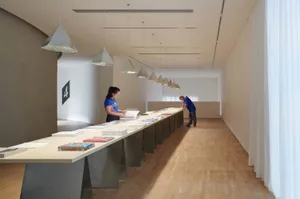 A spacious library with a long, modern table covered in books and papers. People are working and organizing the content.