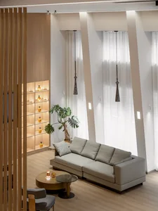 A minimalist living room with a beige sofa, glass coffee table, potted plant, and wooden bookshelf with ambient lighting.