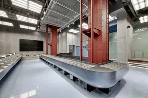 A modern laboratory with exposed concrete walls, a conveyor belt, and red structural columns.