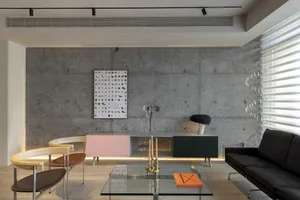 A modern living room with a concrete wall, featuring a glass coffee table, black sofa, and minimalist design elements.