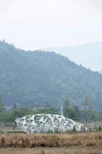 A metallic, modern structure resembling a wave or organic form, located in a field with a mountainous backdrop.