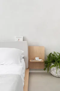 A minimalist bedroom with a white bed, a floating wooden nightstand, and a potted fern plant.