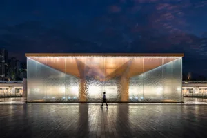 A modern public space featuring a glass water feature illuminated at night with a person walking in front.