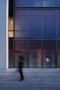 Two blurred pedestrians walking past a modern building with a glass facade reflecting the surrounding environment.