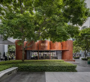 A modern restaurant exterior featuring a metal corrugated facade under a lush tree canopy.