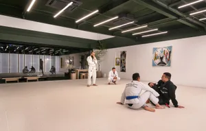 Judo practitioners in white mats sitting and standing in a spacious, well-lit training room with white walls.