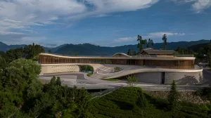 A modern hotel with concrete and glass exterior, surrounded by greenery and mountains.