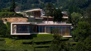Modern residential house with concrete exterior, surrounded by greenery and hills in the background.