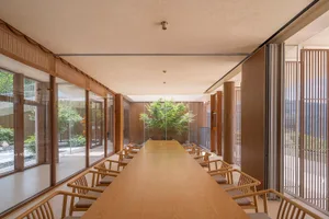 A minimalist cafeteria featuring wooden and glass elements, with a long wooden table and chairs.