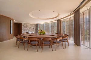 A modern dining room featuring a circular wooden table surrounded by chairs, with a stone wall backdrop and large windows.