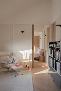 A minimalist living room with white wood furniture and a white rug, featuring a comfortable seating area and a bookshelf.