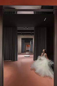 A woman dressed in a flowing tulle dress standing on a pink terrazzo floor, with dark curtains and minimalistic lighting in t