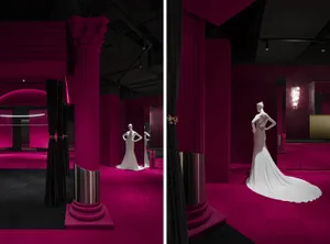 A pink exhibition space with a mannequin wearing a long white dress on a platform, surrounded by black curtains and a black c
