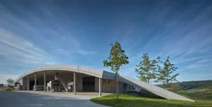 Modern, curved, concrete winery building with a sloping ramp and green surroundings.