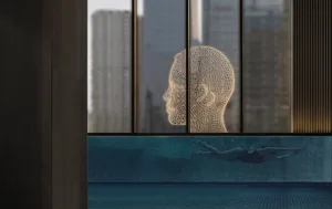 A metallic grid sculpture in the shape of a human head is displayed behind a window pane.