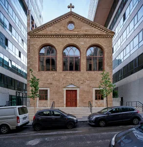 A modern stone church with three arched windows and a cross on the roof, surrounded by modern buildings and parked cars.