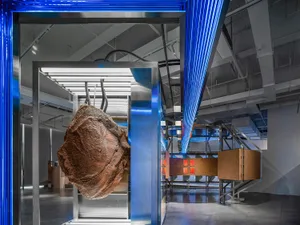 A modern installation art piece featuring a metallic sculpture suspended in a metallic-framed enclosure with blue light beams