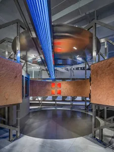 A modern museum exhibition space featuring a reflective metal installation with blue neon lights and red illuminated panels.