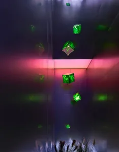 An abstract installation featuring green glass cubes suspended in a dark room with pink and purple lighting.